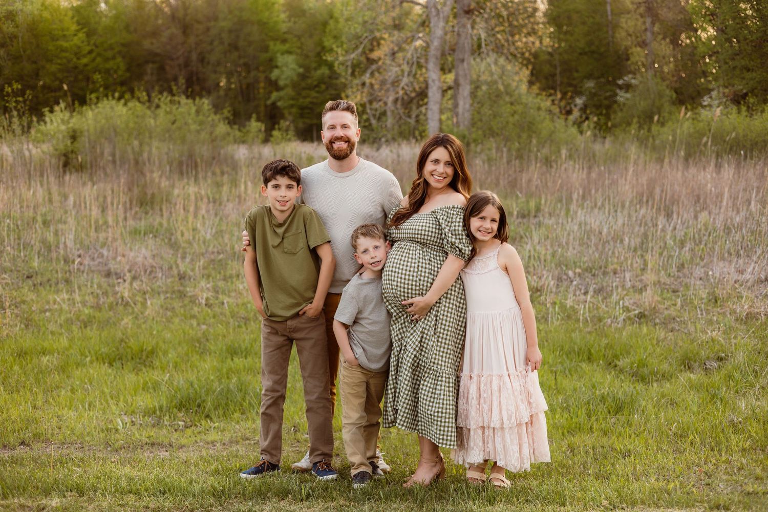 Family of five posing outdoors in green field, pregnant mother in floral dress with husband and three children.