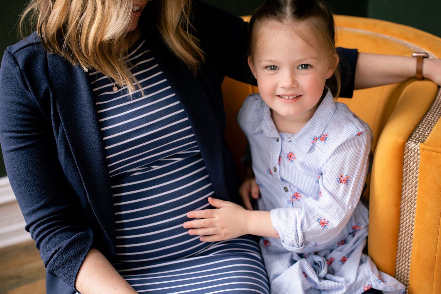 Young girl smiles and touches pregnant mothers belly while sitting together in a yellow chair.