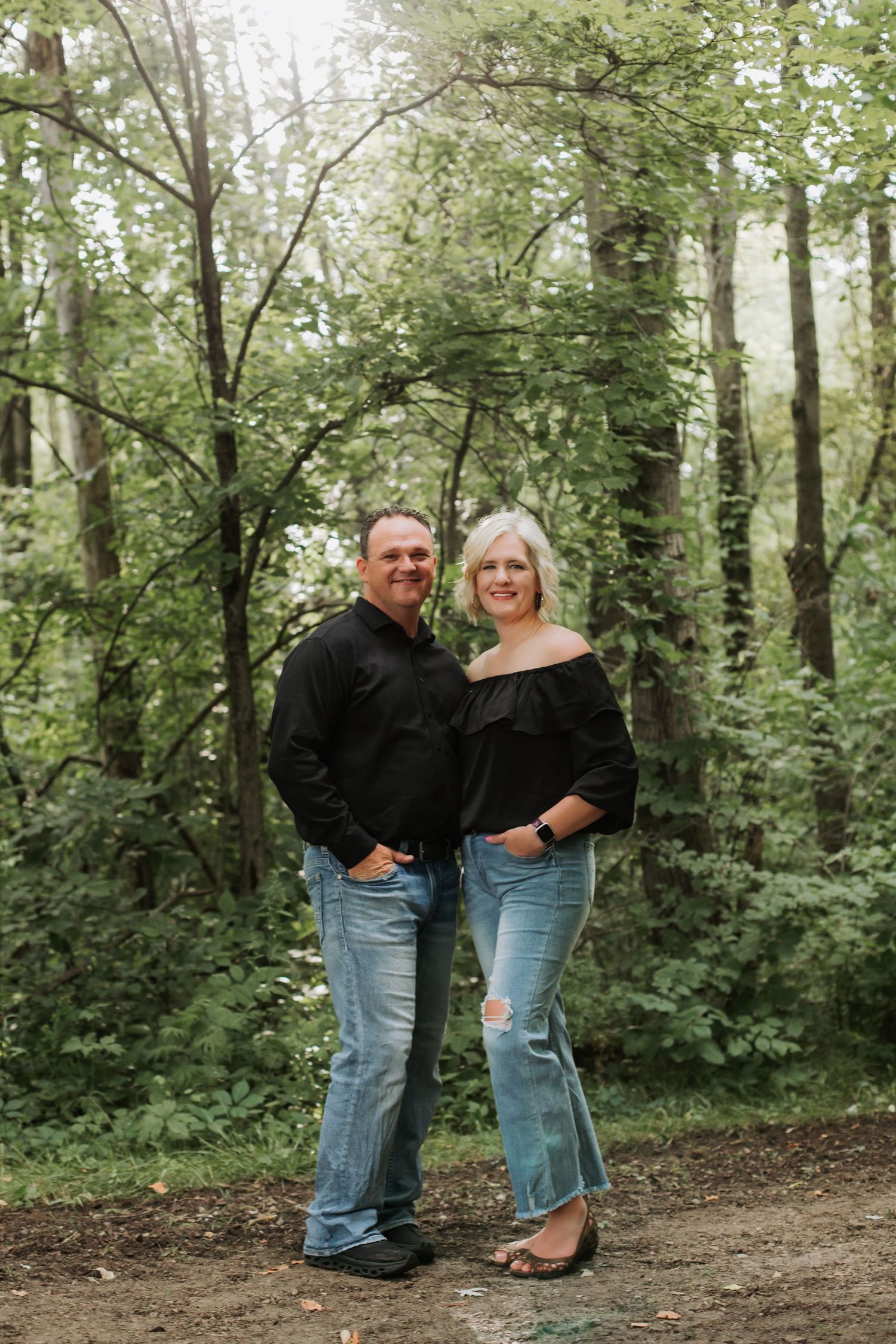 Smiling couple in black tops and jeans standing in a lush green forest.