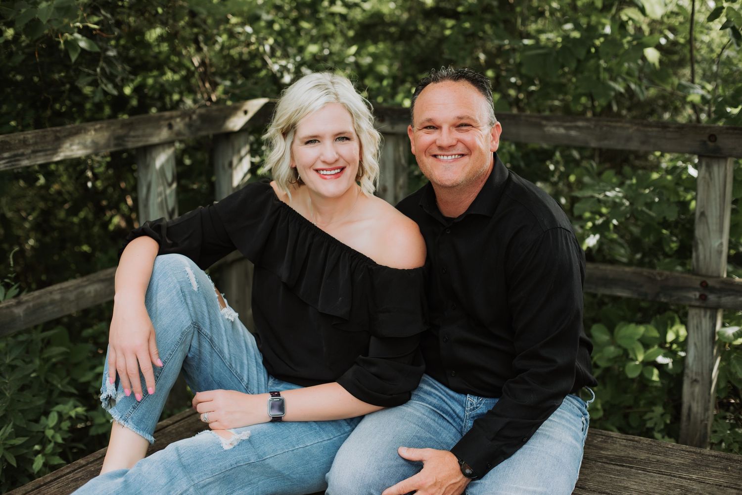 Smiling couple sitting together on a wooden bridge surrounded by lush green foliage.