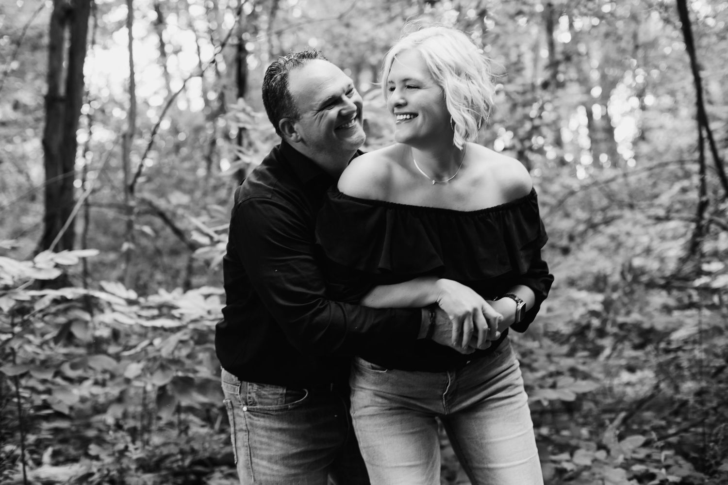 A black and white photo of a couple sharing a tender embrace and smiling in an autumn forest.
