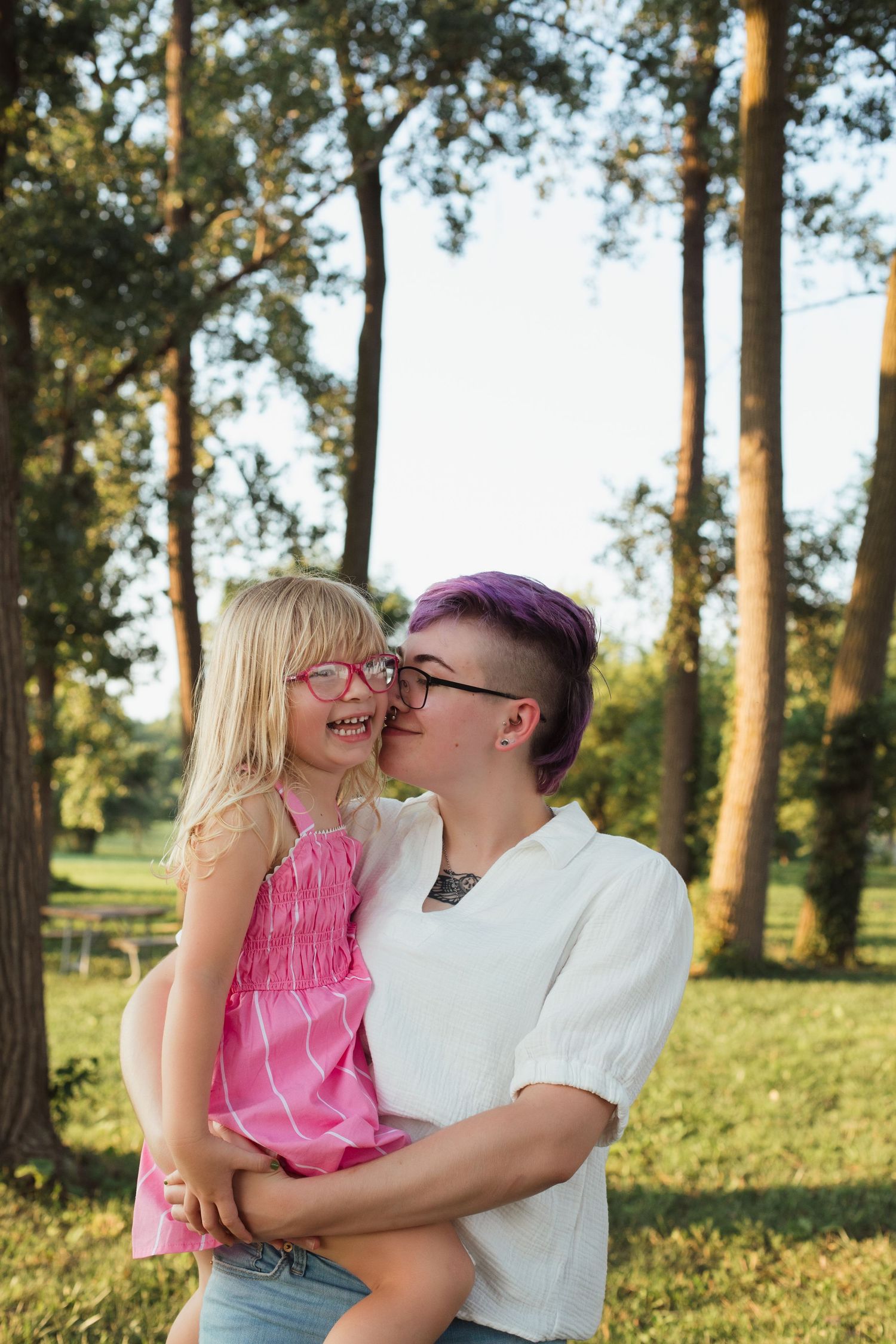 Woman with purple hair kissing smiling blonde girl in pink dress outdoors in park.