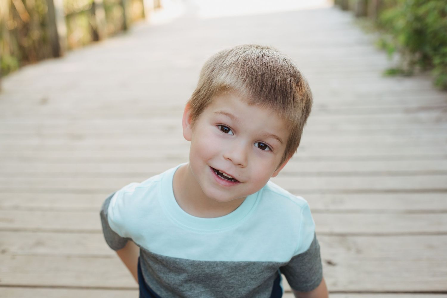 Cute young boy in teal and gray shirt smiles candidly on sunny wooden boardwalk.