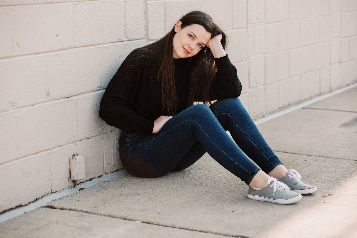 Young woman in black sweater and jeans sitting against a gray wall outdoors, smiling casually.