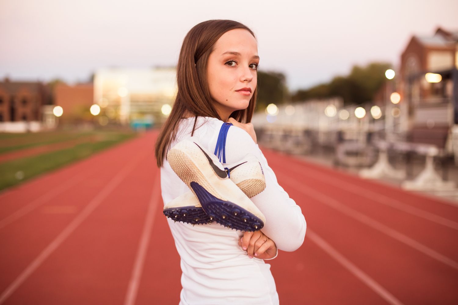 Young female athlete in white Nike top holds blue track spikes on a red running track at dusk.