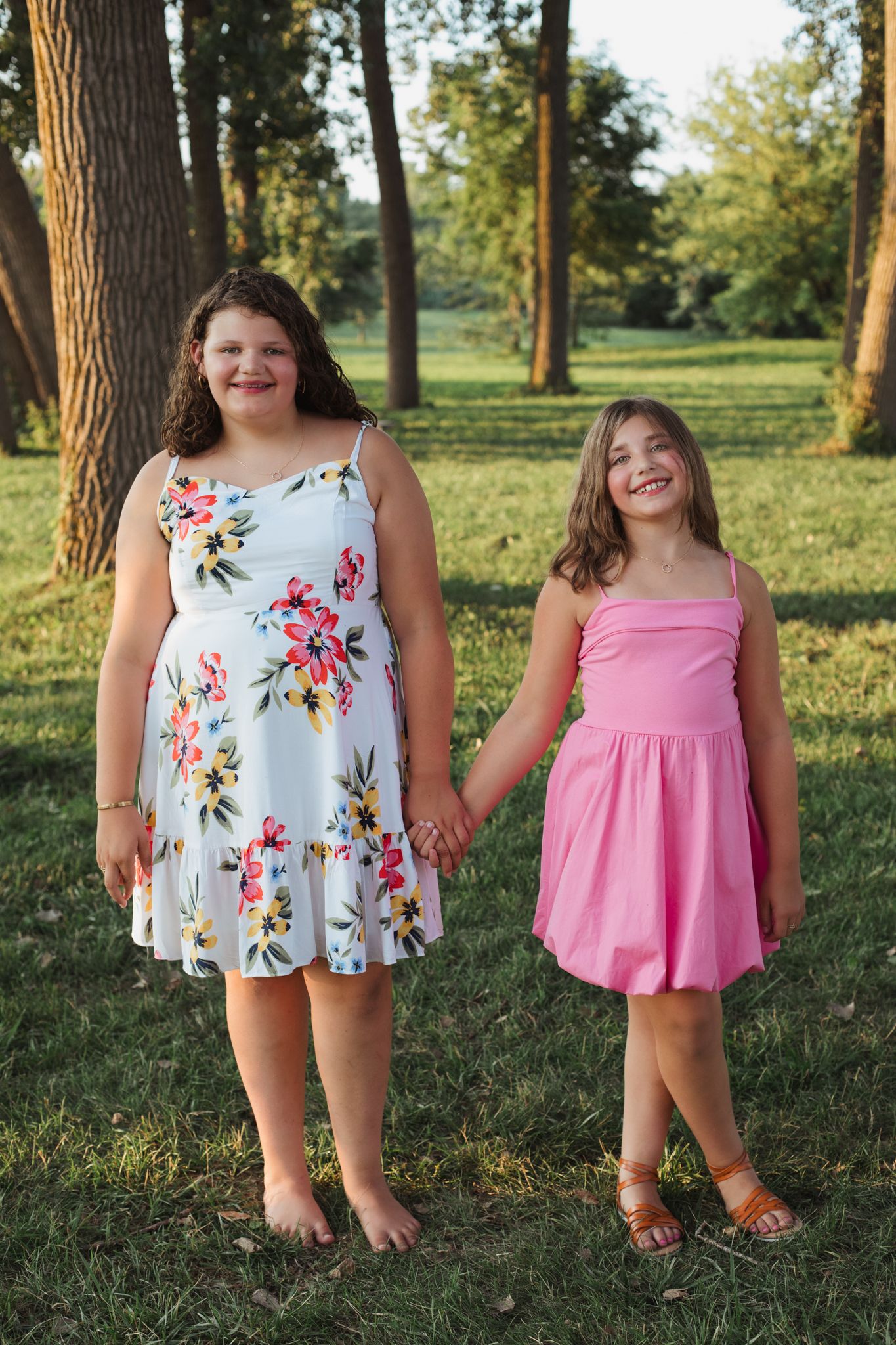 Two girls in floral and pink dresses hold hands smiling in a sunny park.