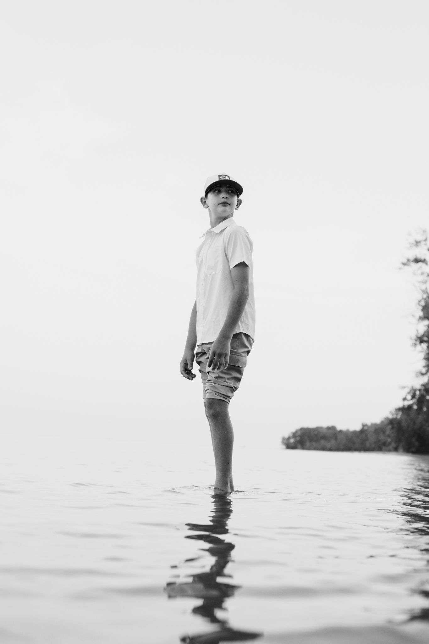Black and white photo of a young man standing in shallow water wearing a cap, shorts, and t-shirt.