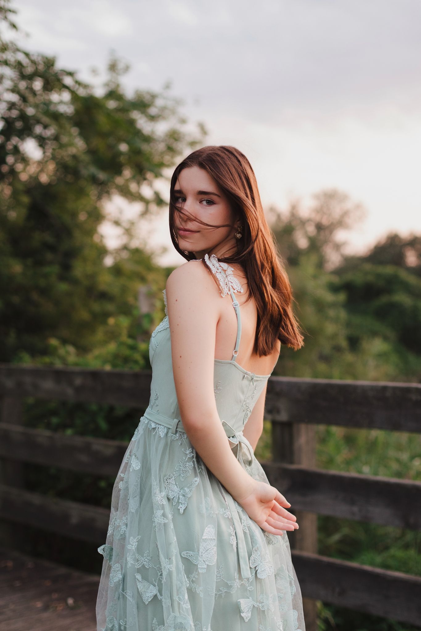 Young brunette woman in mint green butterfly dress turns back to camera on wooden bridge.