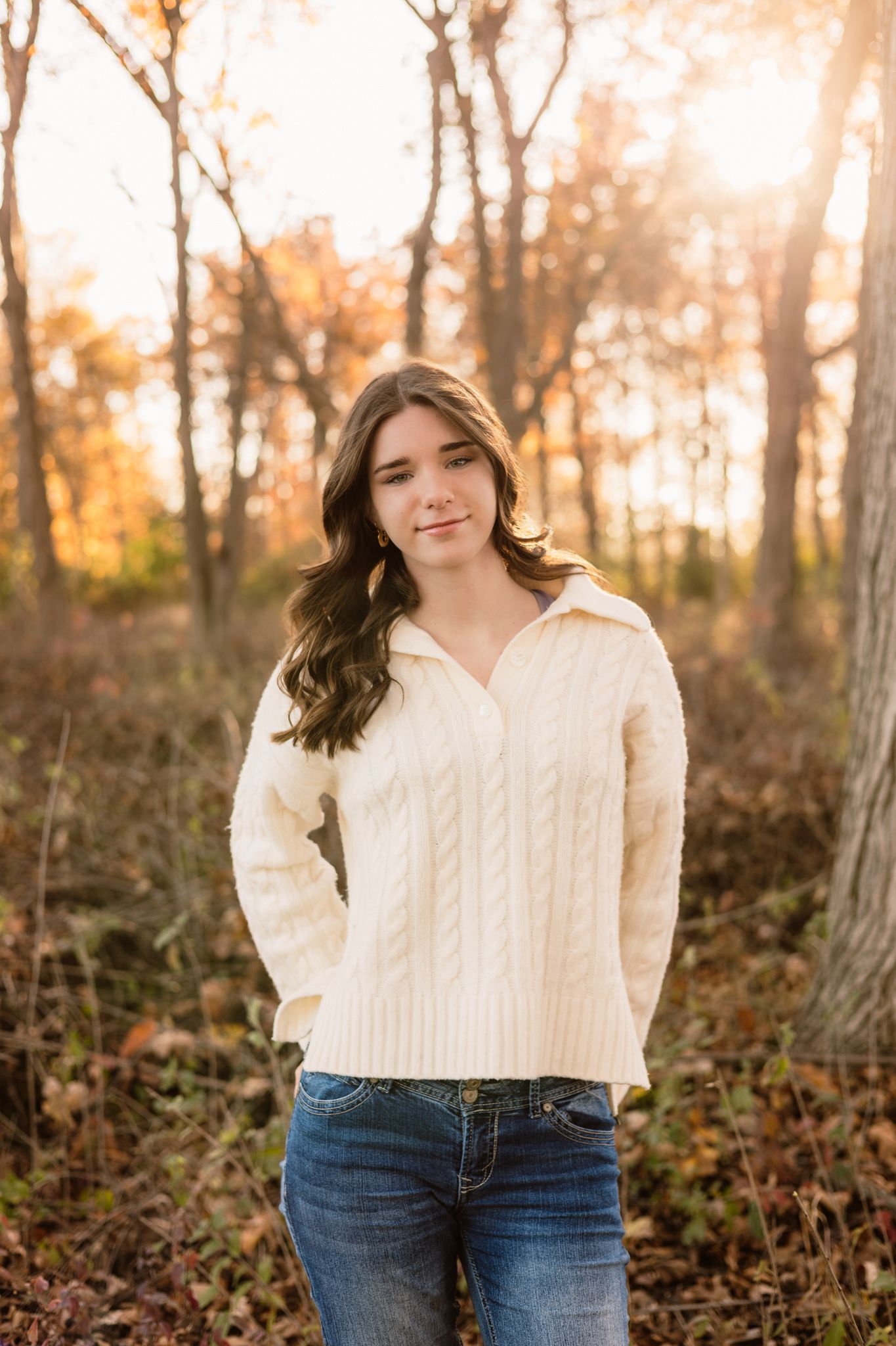 Young woman in cream cable-knit sweater and jeans standing in autumn forest at sunset.