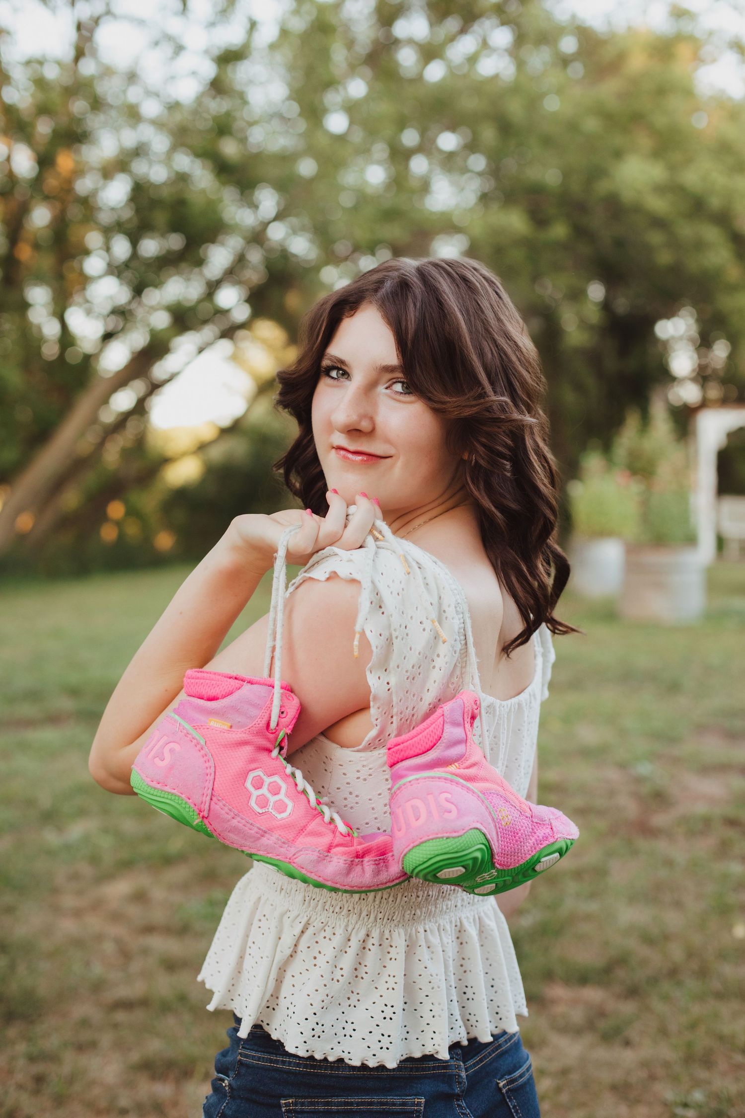 Young woman in white lace top holding pink cowboy boots outdoors at sunset.