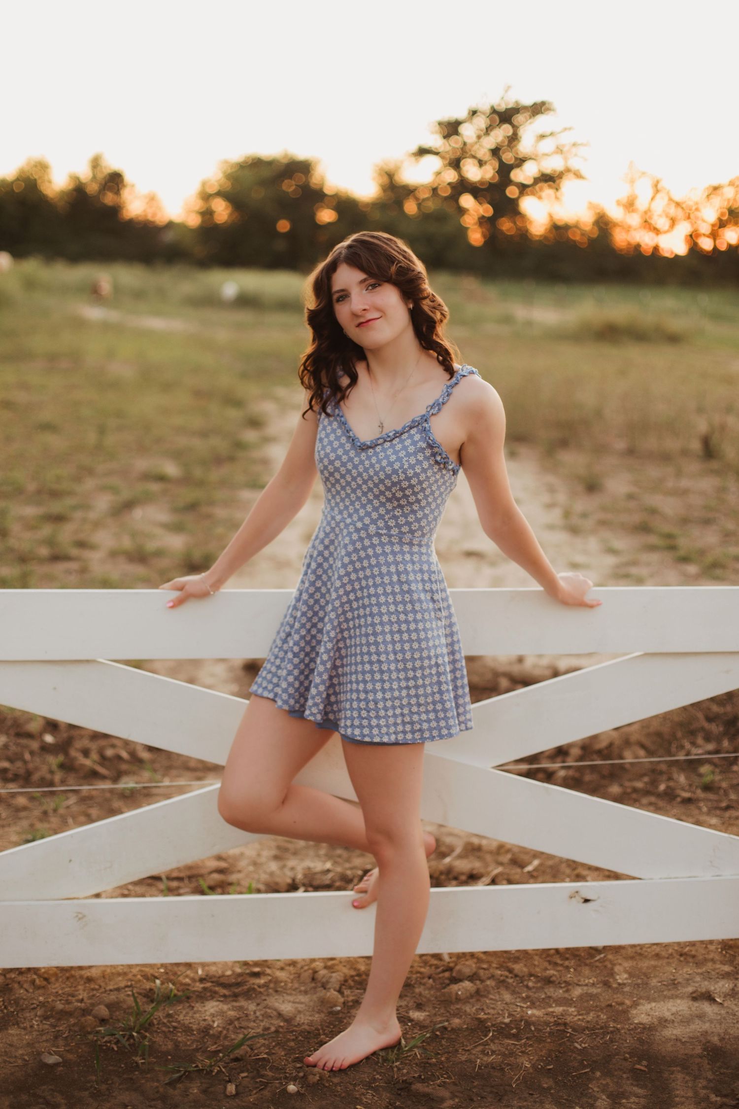 Young woman in blue gingham dress leaning against a white wooden fence at golden hour sunset.