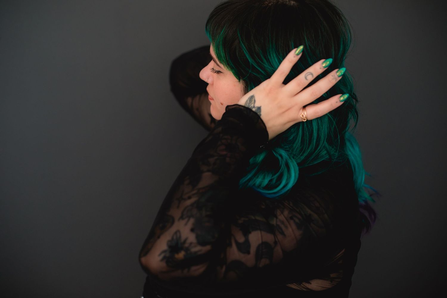 Woman with teal green hair in black floral lace top posing with hands on head against gray wall.