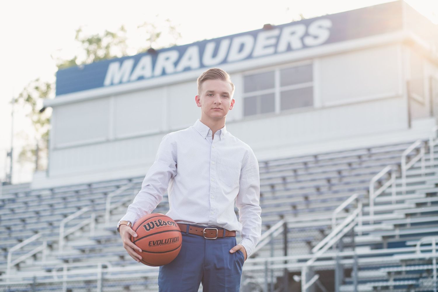 Teen boy in white dress shirt holds basketball at Marauders stadium bleachers for senior portrait photo.