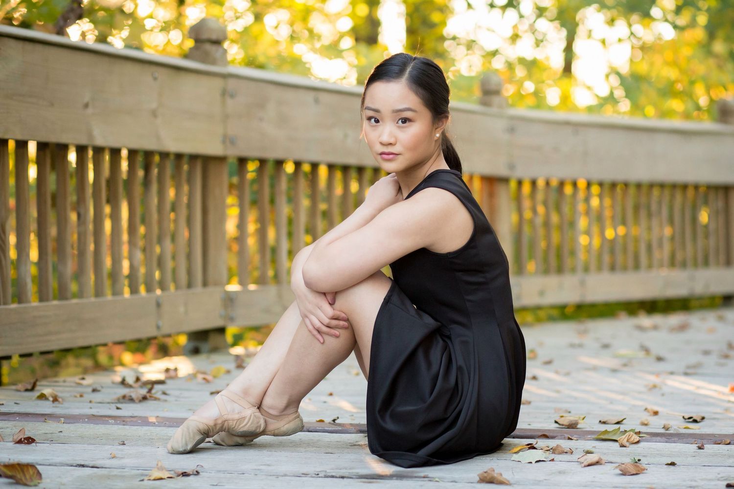 Young ballet dancer in black dress and pointe shoes sits gracefully on wooden deck surrounded by autumn leaves.