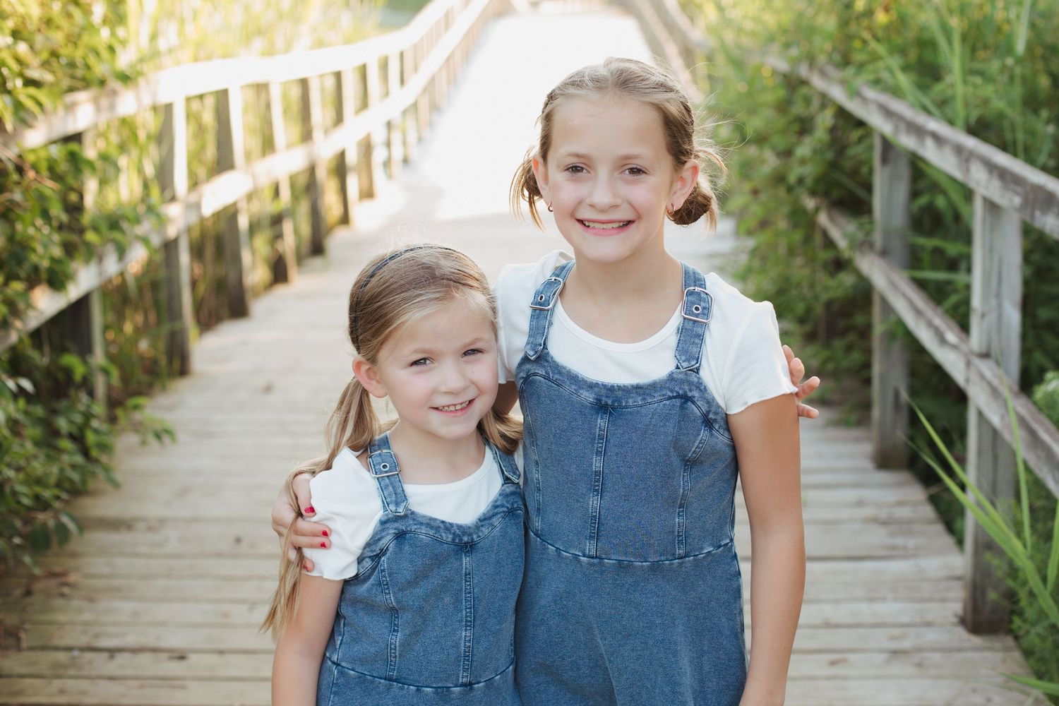 Two sisters in matching denim overalls smiling on a wooden boardwalk outdoors.