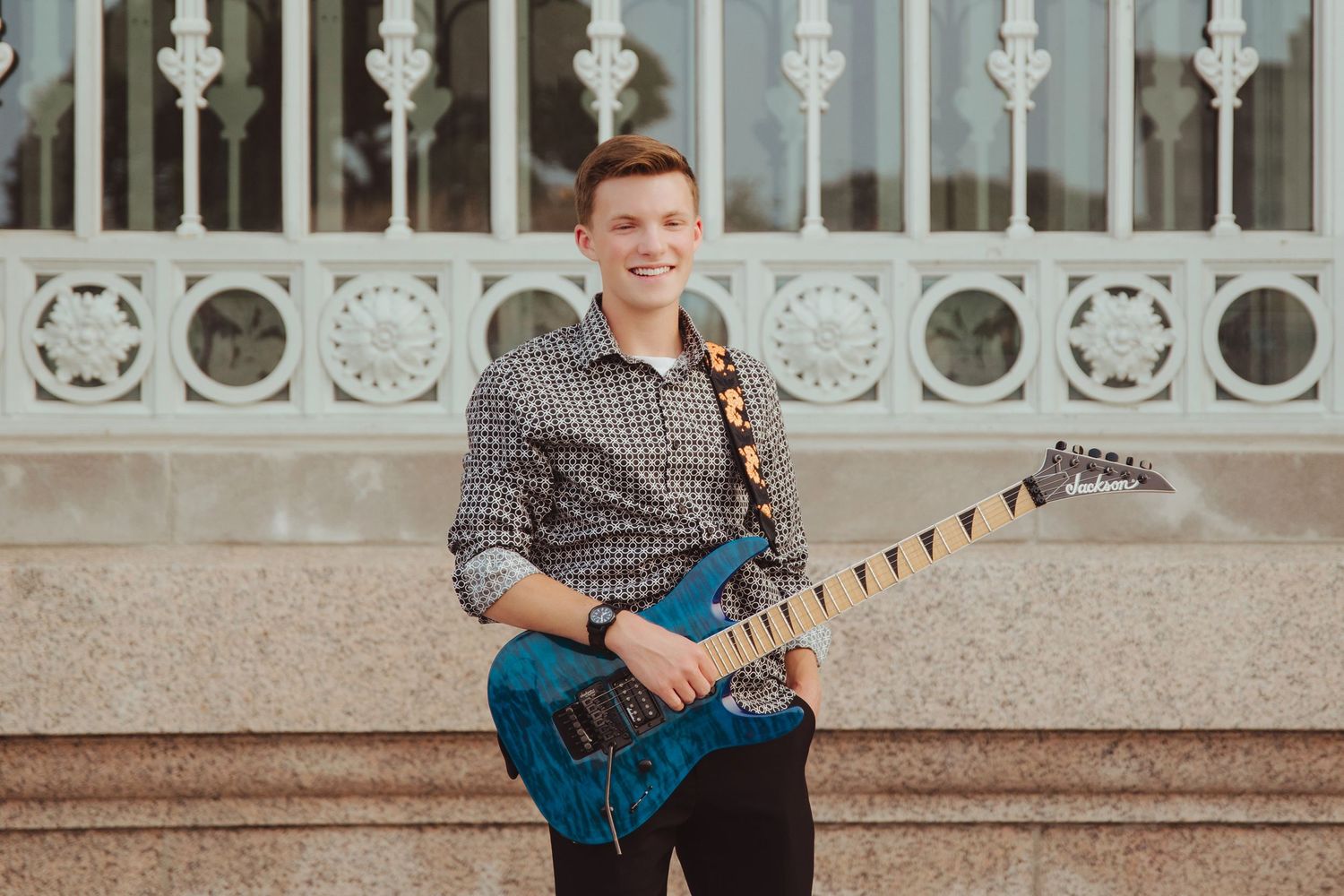 Young man smiling with blue electric guitar outdoors near ornate building.