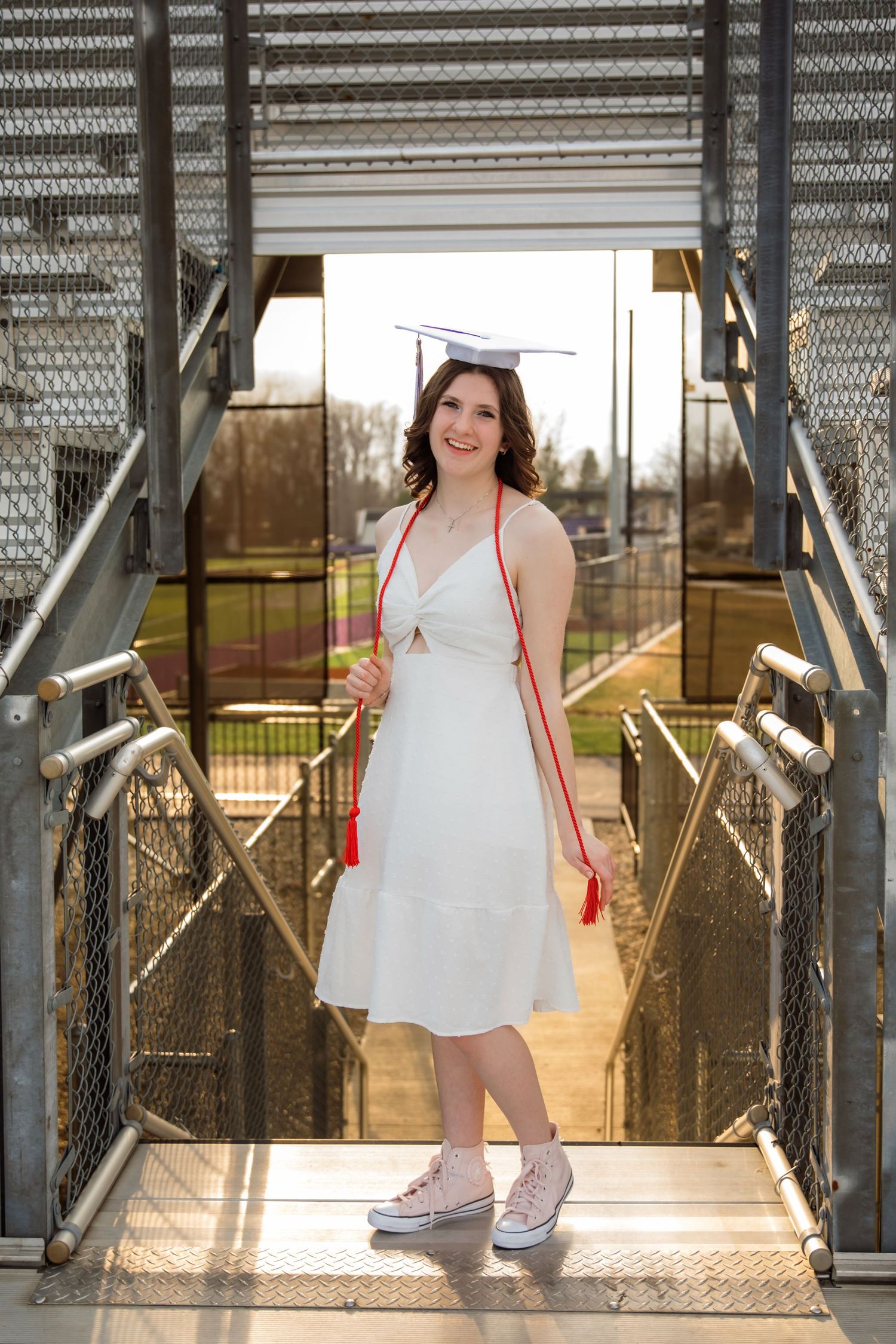 A smiling graduate in white dress and cap holds red tassel on metal walkway outdoors.