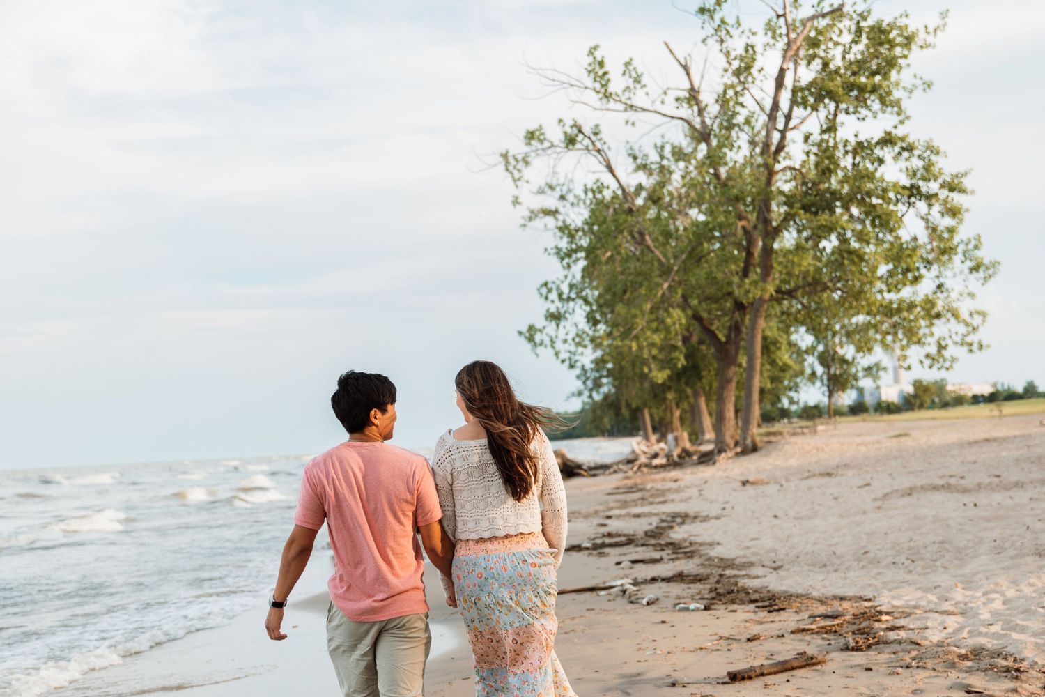 Couple walking hand in hand along a sandy beach shoreline with trees in the background.