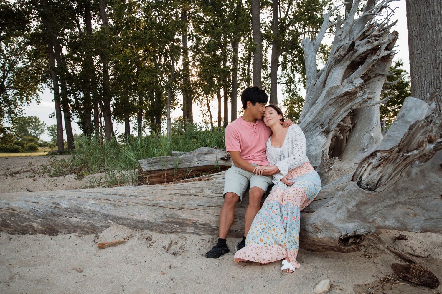 Romantic couple sitting together on driftwood log at sandy beach surrounded by tall trees at sunset.