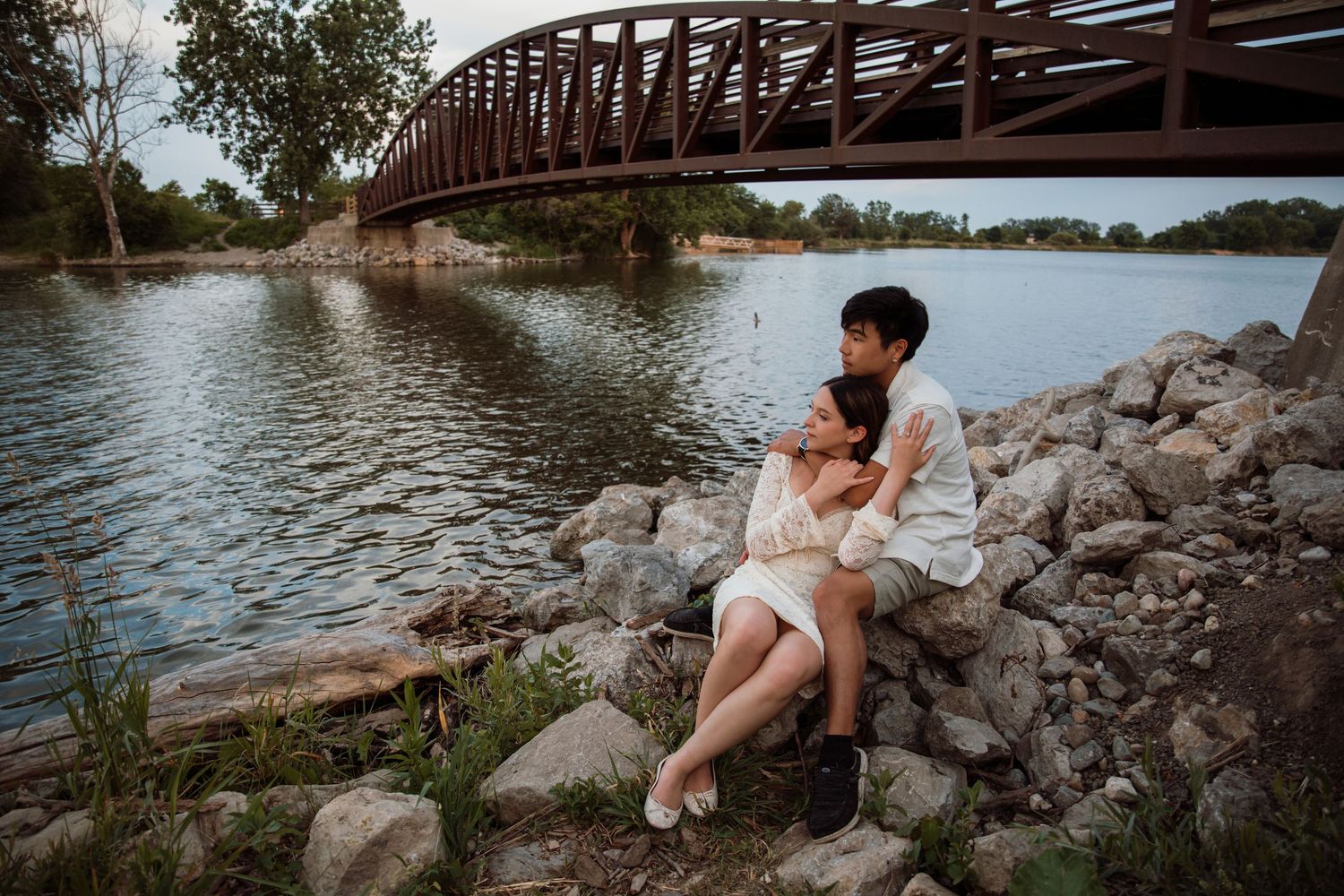 Couple sitting closely on rocks by a river, embracing under a wooden bridge at dusk.