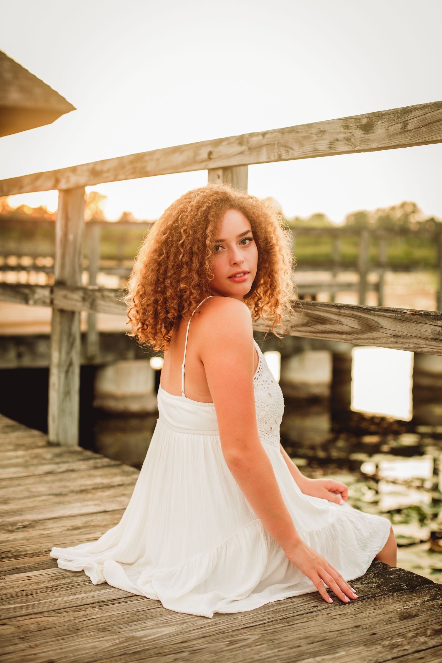 Young woman with curly red hair in white dress sitting on wooden dock at sunset.