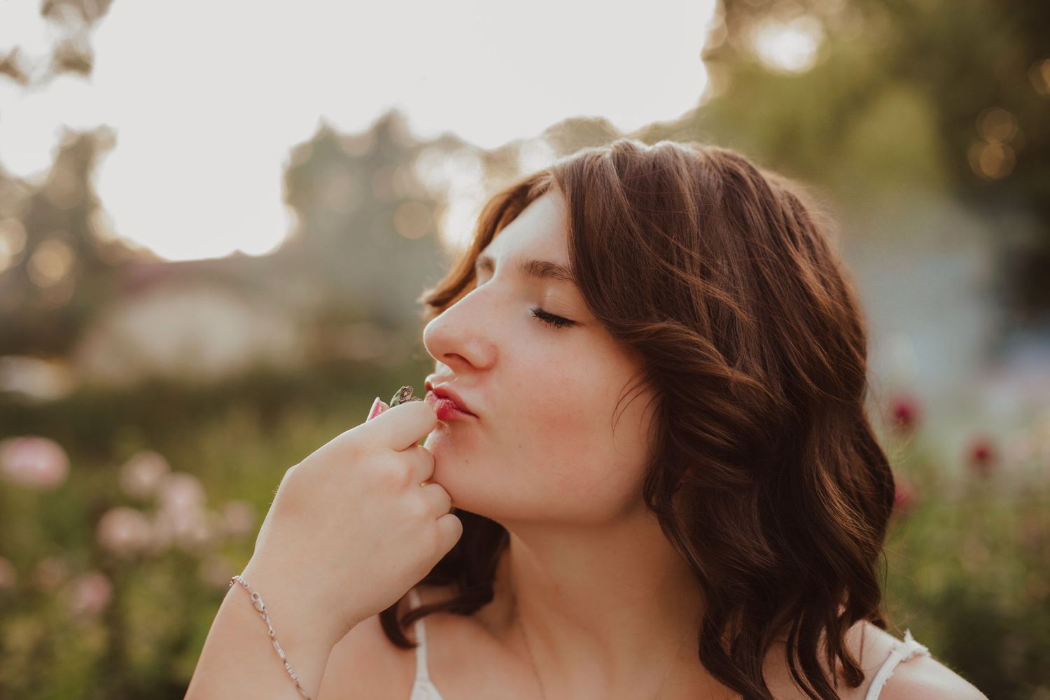 Young brunette woman smelling a small white flower with eyes closed in a sunlit garden.