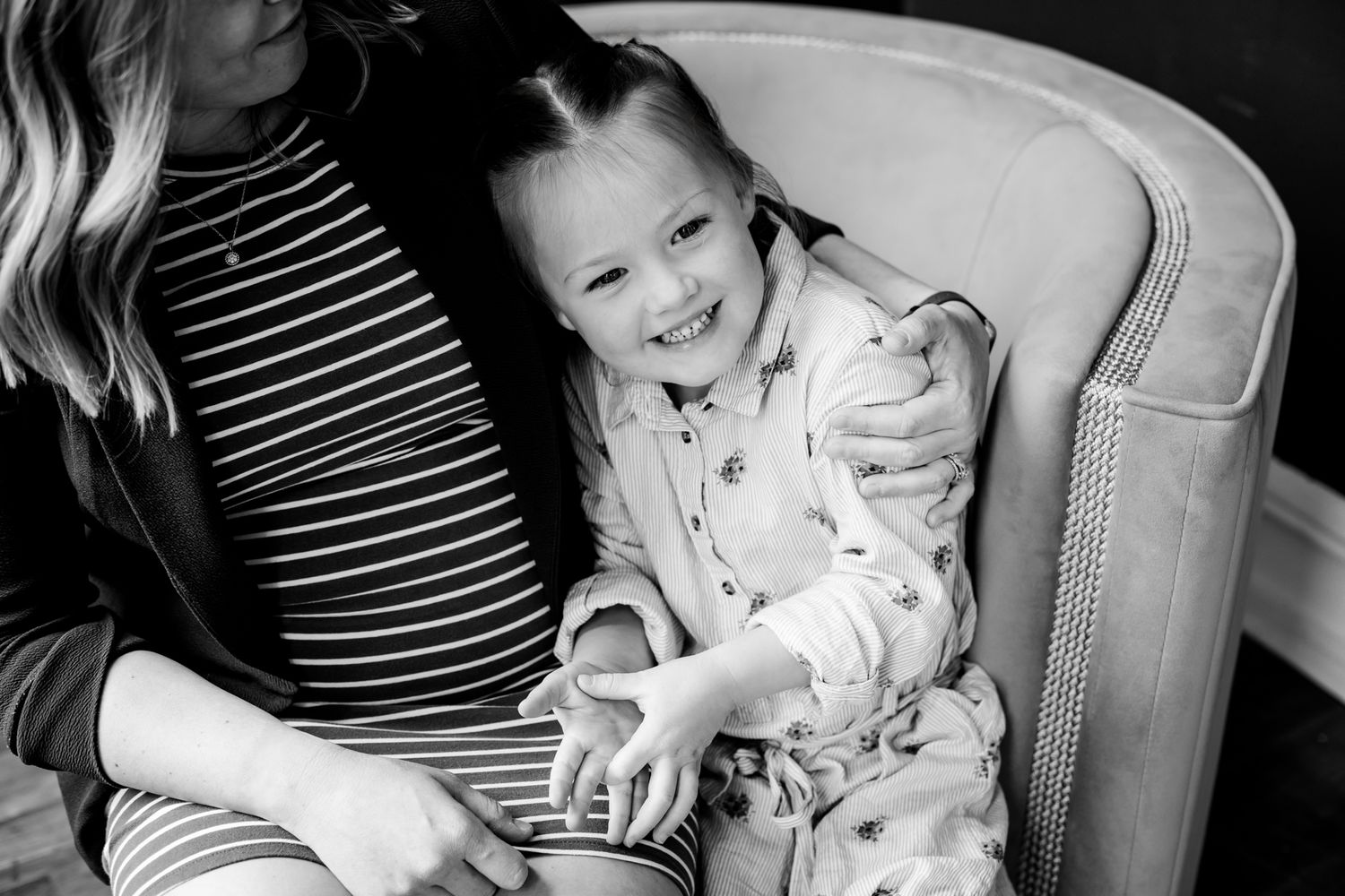 Smiling young girl sits on a chair beside a pregnant woman in a black and white maternity photo.