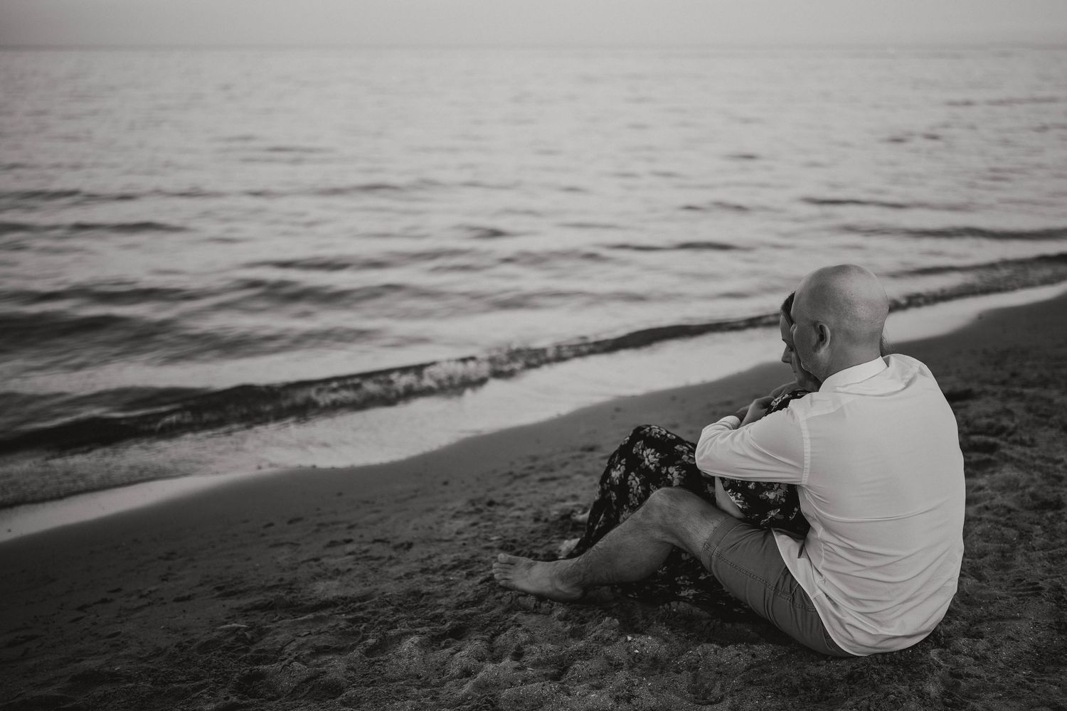 Black and white photo of a bald man sitting alone on a sandy beach at sunset.