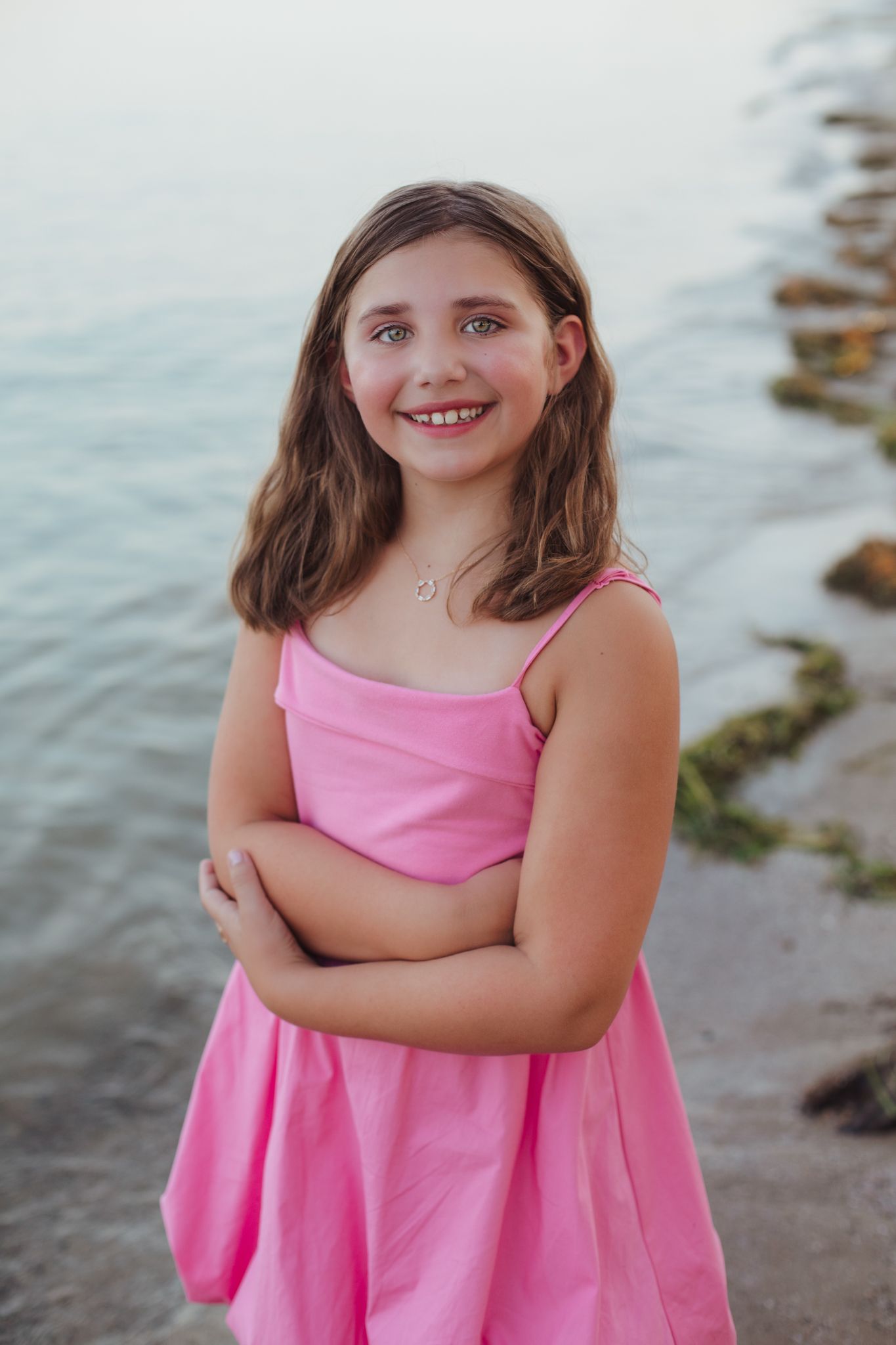 Smiling young girl in a pink dress stands by the water with arms crossed at the beach.