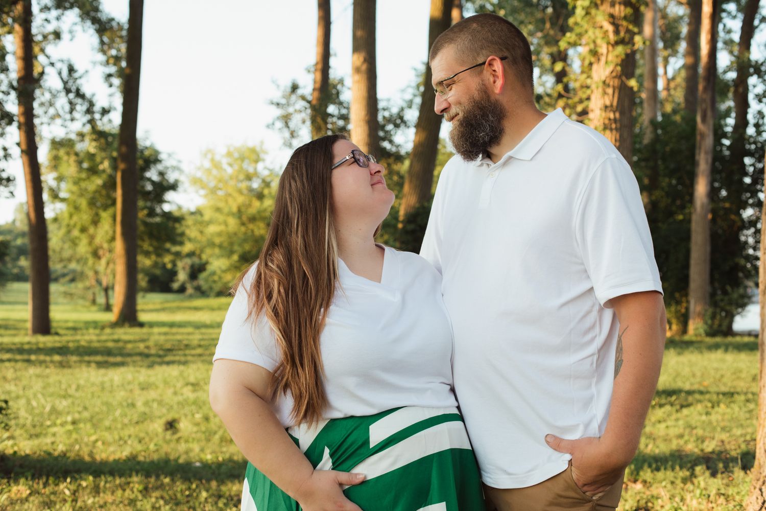 Couple smiling at each other in a sunny park, woman in white top and green skirt, man in white polo shirt.