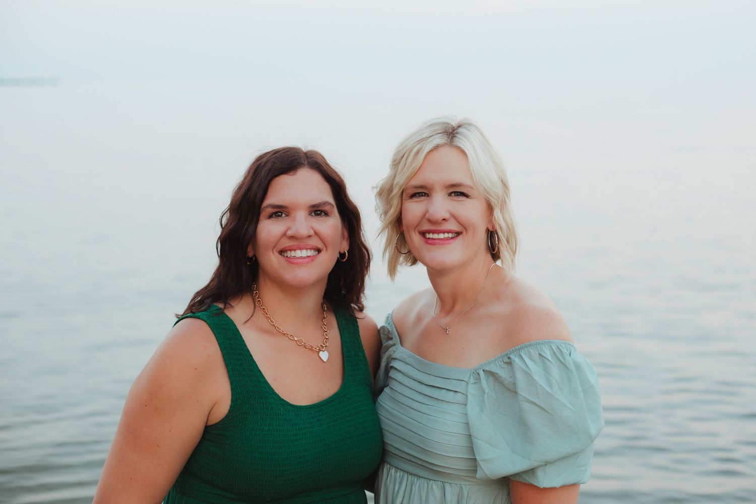 Two smiling women pose together near water, one in green dress, one in light blue off-shoulder dress.