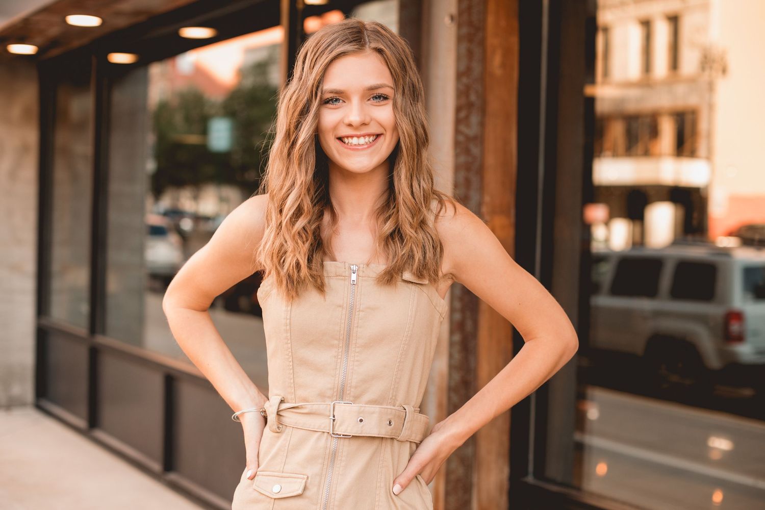 Young woman in beige belted jumpsuit smiling outdoors on urban street with buildings in background.