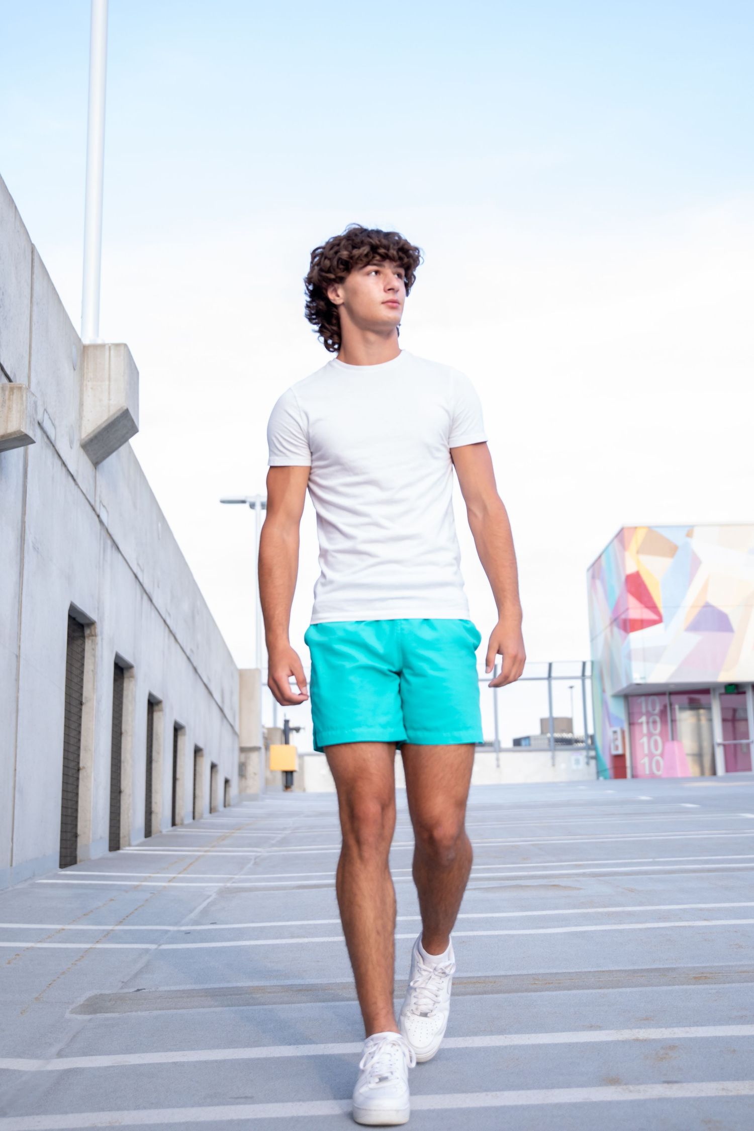Young man with curly hair walks outdoors in white t-shirt, turquoise shorts, and white sneakers against urban backdrop.