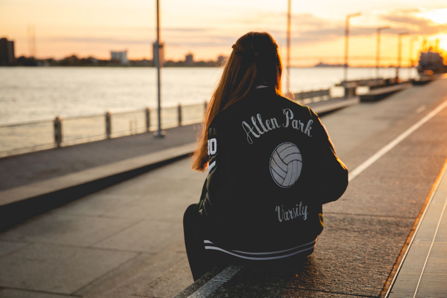Girl wearing Allen Park Varsity volleyball jacket sitting by waterfront at sunset.