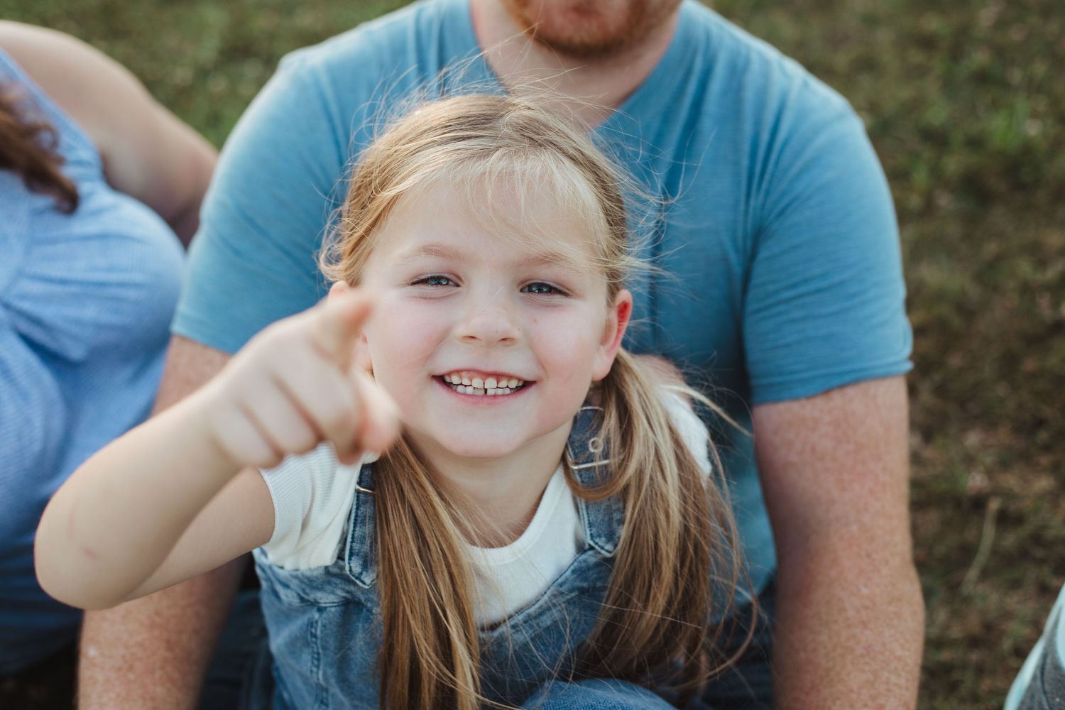 Smiling young girl with pigtails pointing at camera while sitting outdoors in grass.