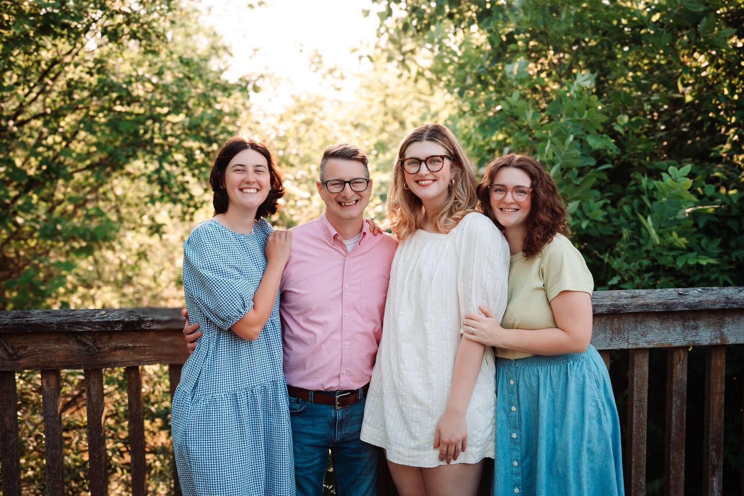 Father poses with three daughters on a sunny wooden deck with green trees in the background.