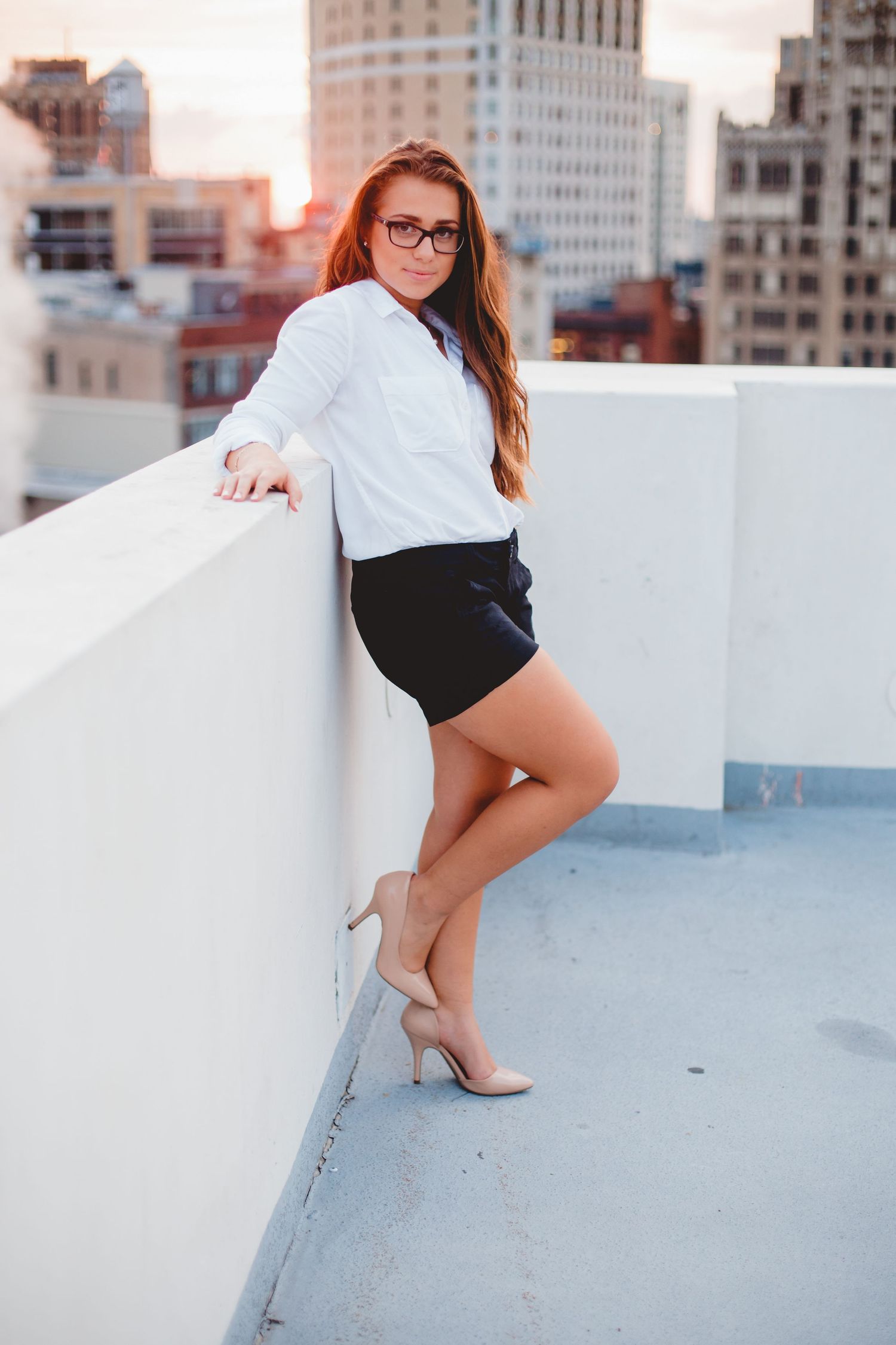 Young woman in white shirt and black shorts poses on rooftop with city skyline at sunset.