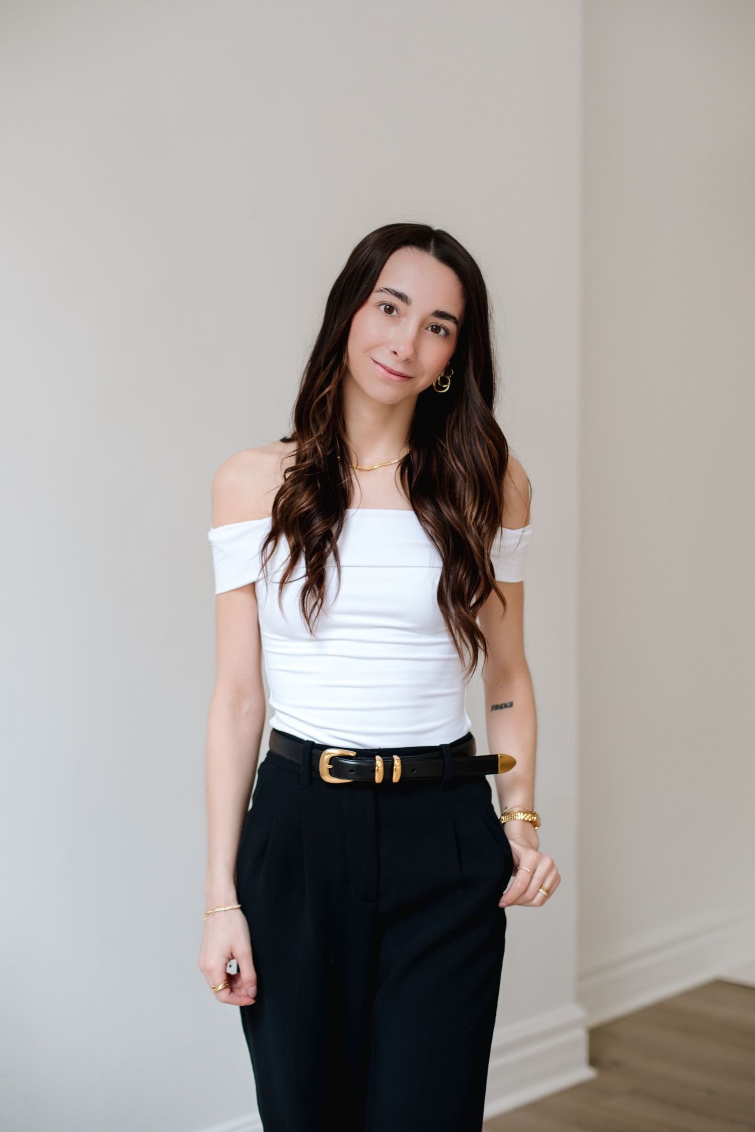 Young woman in white off-shoulder top and black trousers with gold belt posing against neutral wall.
