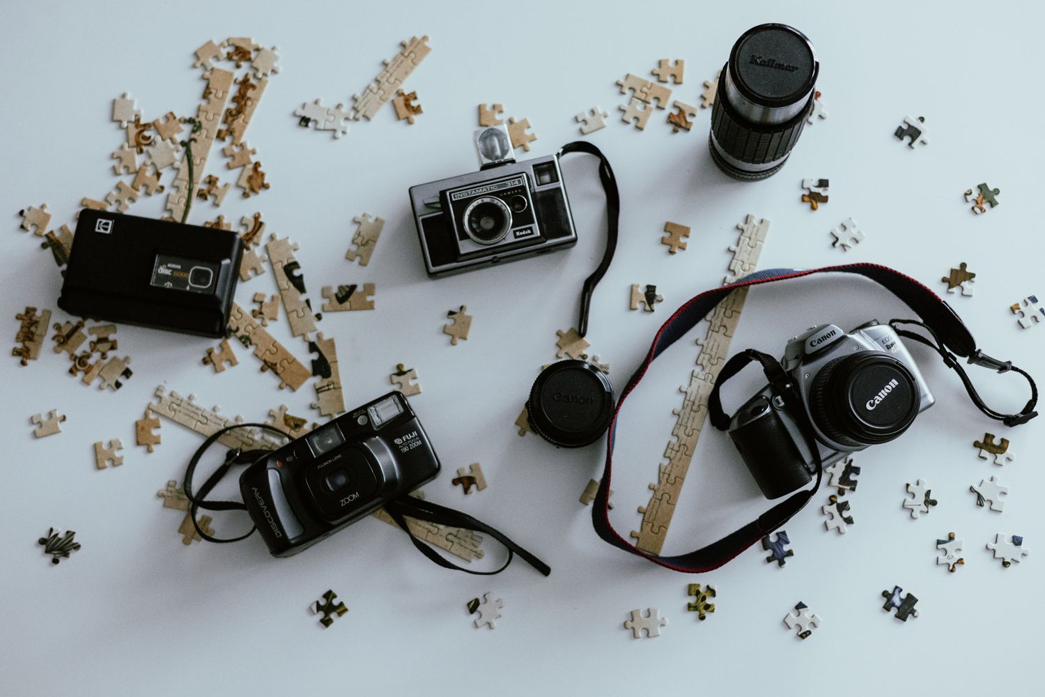 Flat lay of vintage cameras and lenses scattered among puzzle pieces on a white surface.
