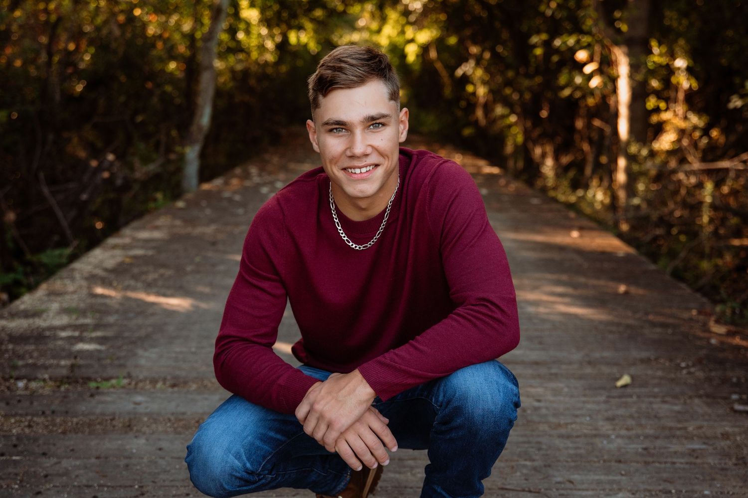 A smiling young man in a burgundy sweater crouching on an outdoor pathway surrounded by colorful autumn trees.