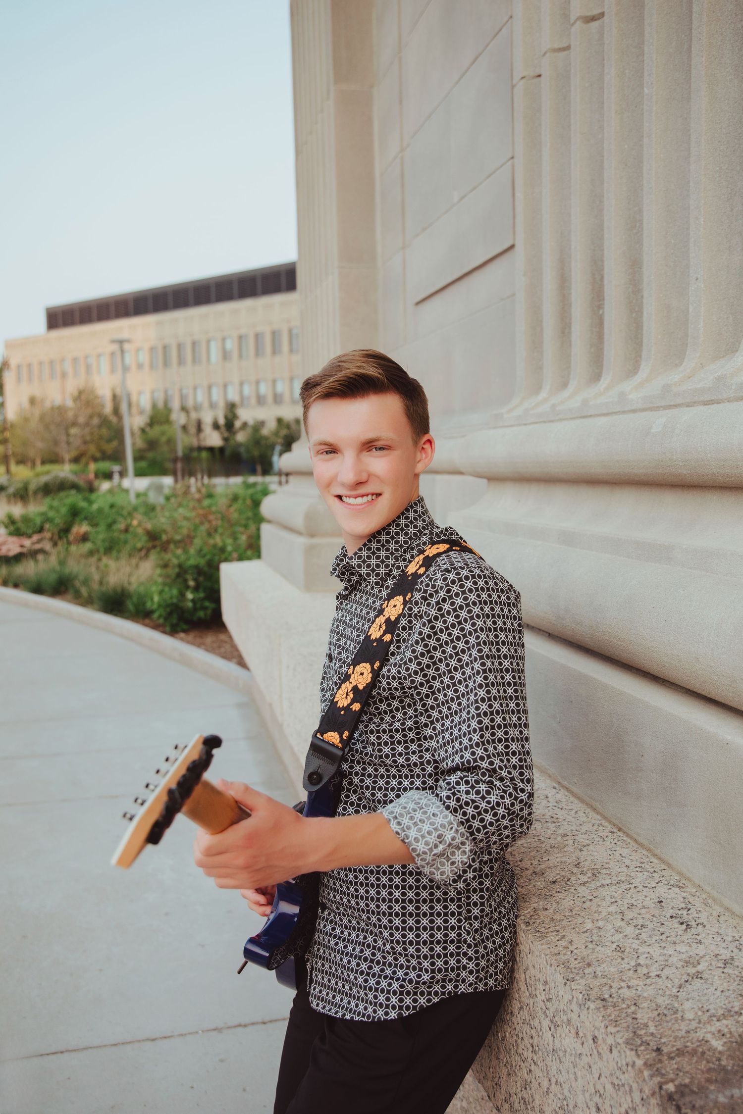 Young man smiling while holding a blue electric guitar with floral strap against stone column outdoors.
