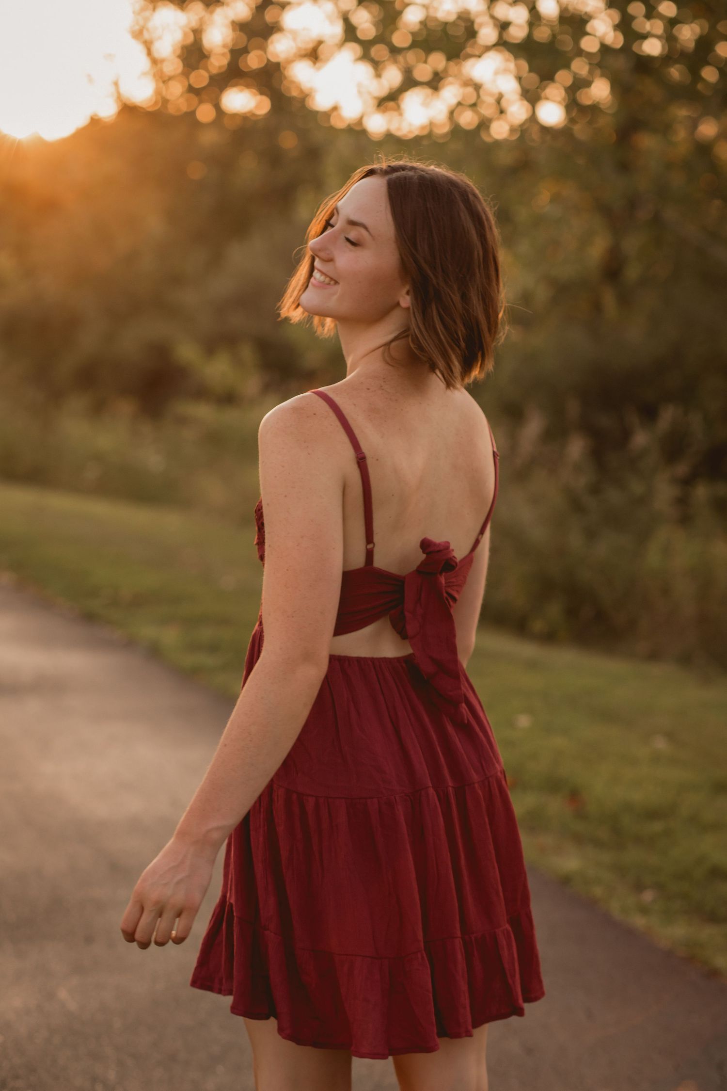 Young woman in backless burgundy mini dress smiling during golden hour on outdoor path.