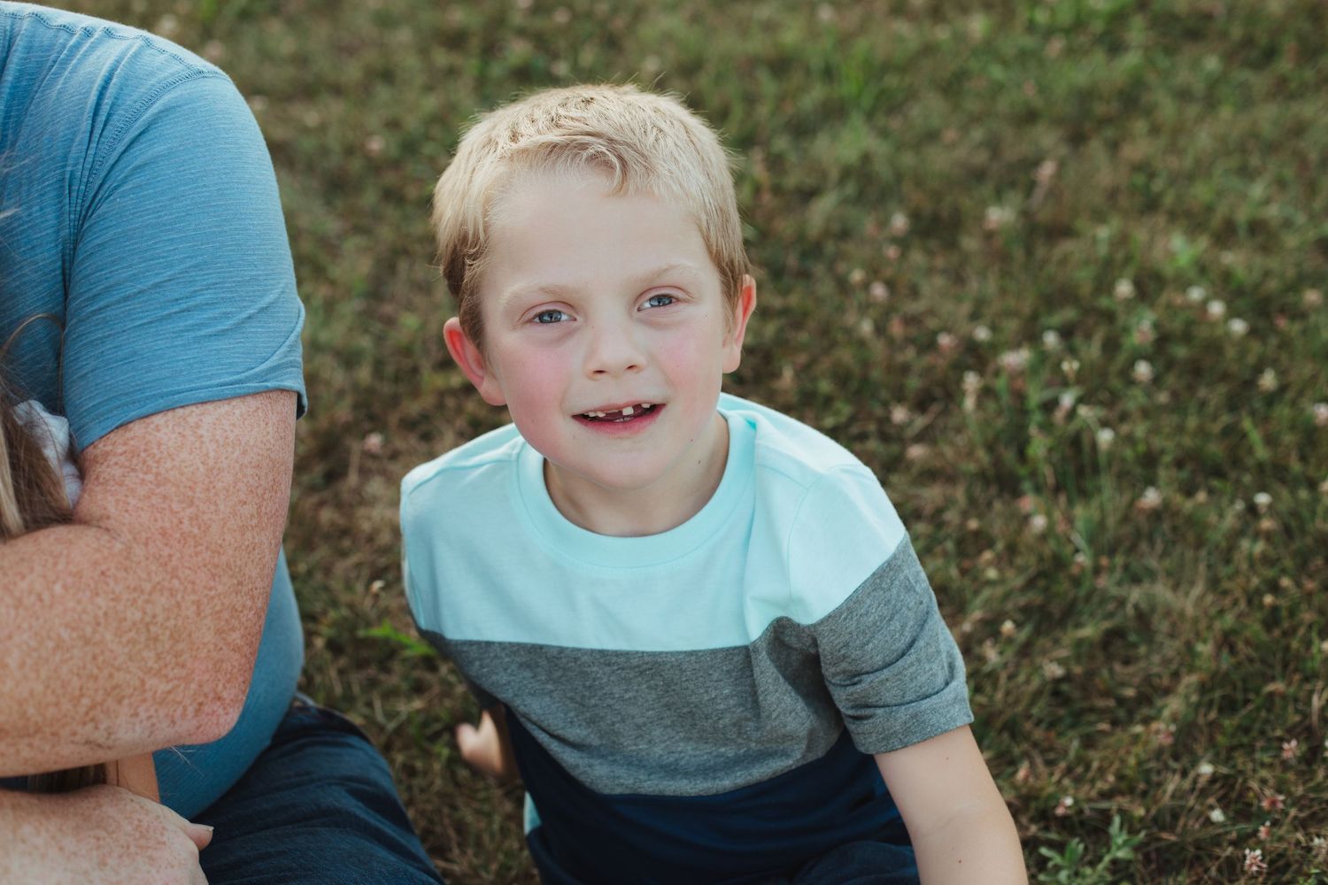 A blonde boy in a teal and gray striped shirt smiles while sitting on grass during an outdoor family portrait session.