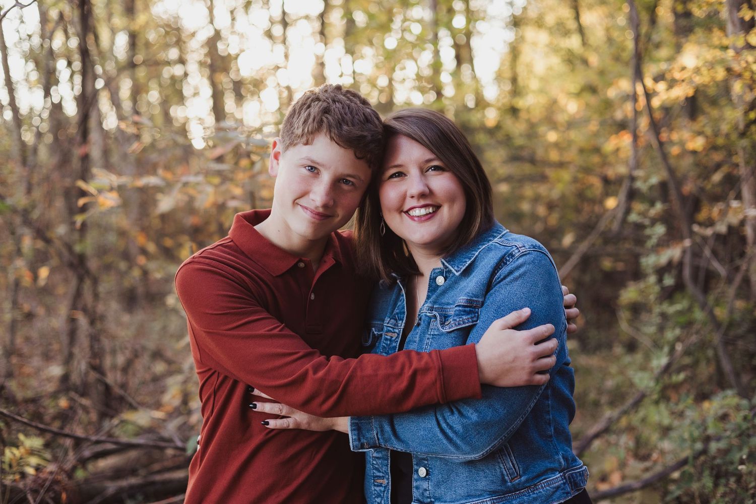 A teenage boy and girl in casual fall outfits hug and smile together in a colorful autumn forest setting.