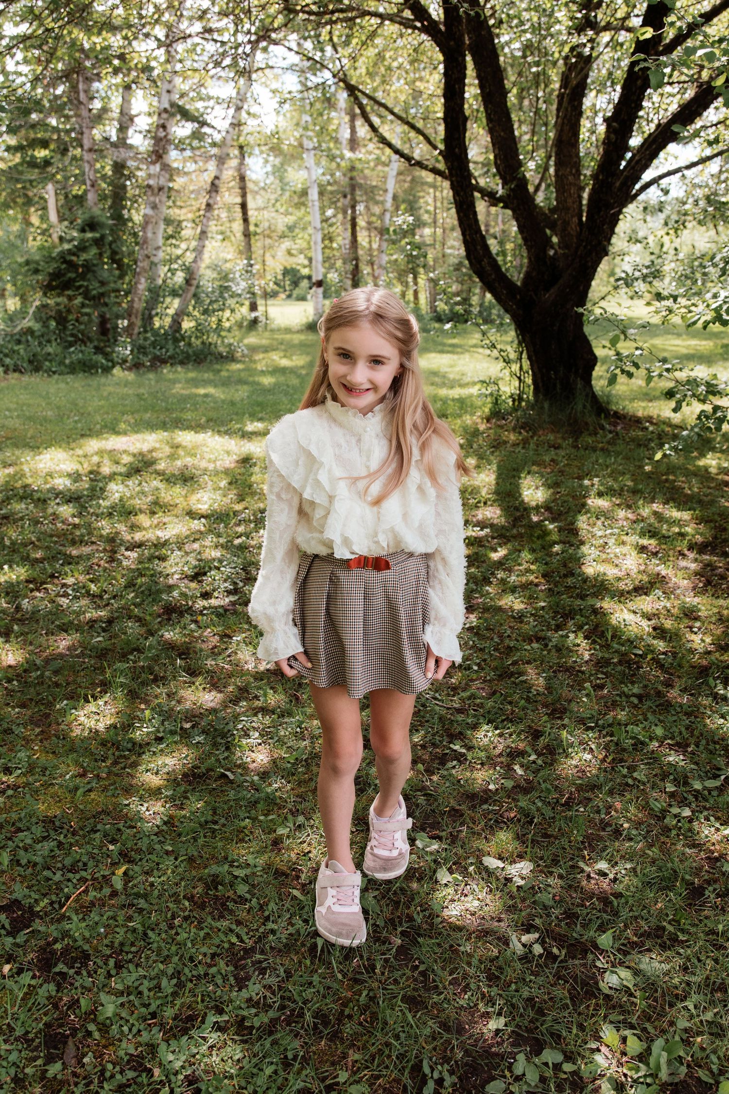 Young girl in white ruffled blouse and plaid skirt smiling outdoors in a lush green park setting.