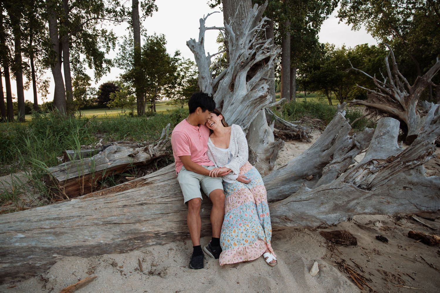 Couple sitting together on driftwood in a sandy park, woman in floral dress leaning on mans shoulder at sunset.