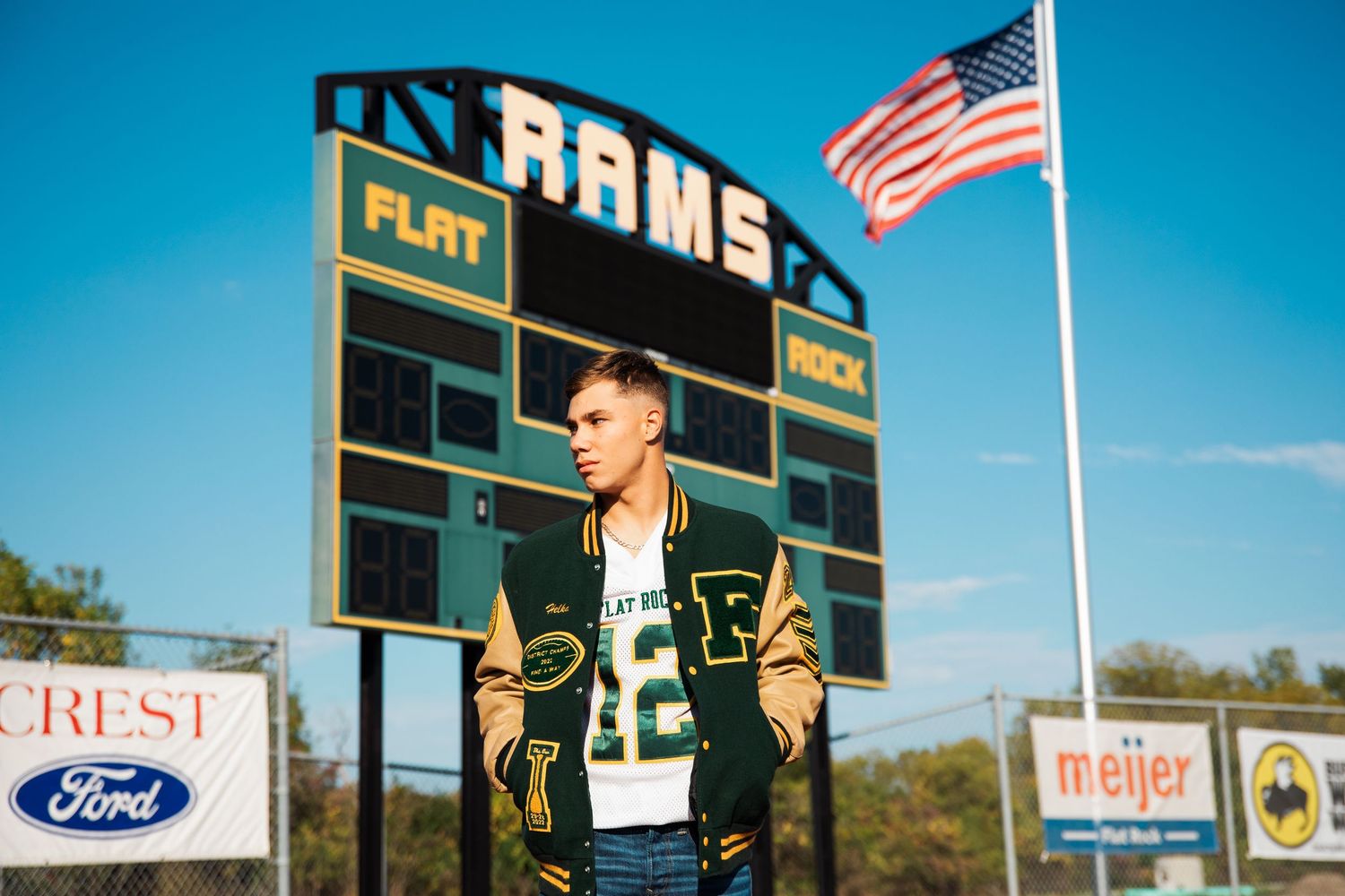 Young man in letterman jacket poses at Flat Rock Rams football field scoreboard with American flag.