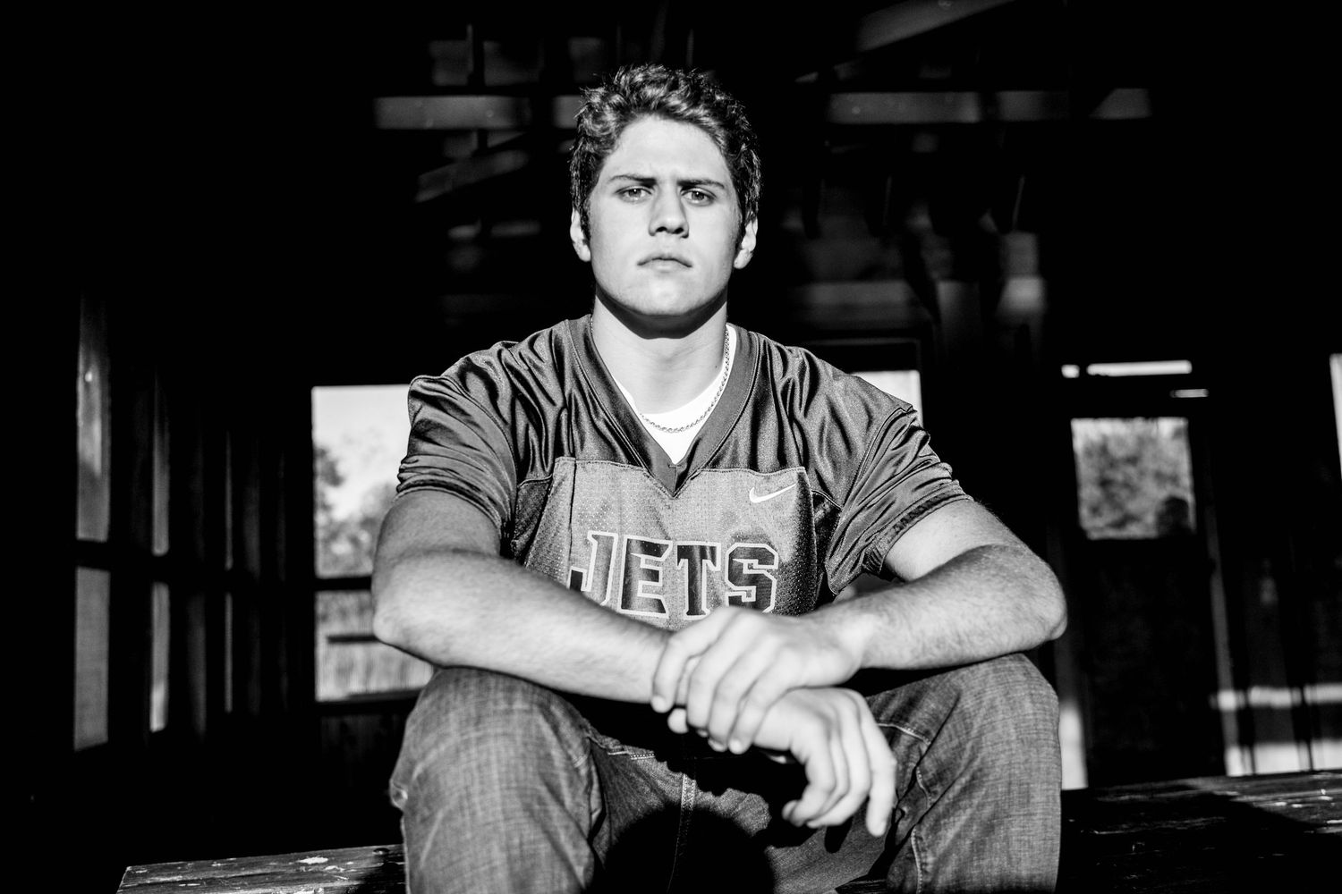 Young man in Jets jersey sits pensively outdoors in black and white portrait photo.