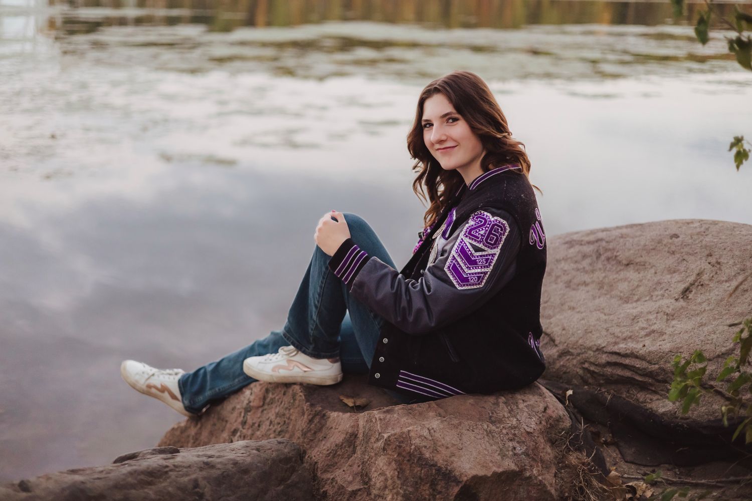 Young woman in varsity jacket sitting on rocks by a lake, smiling for senior portrait photos outdoors.