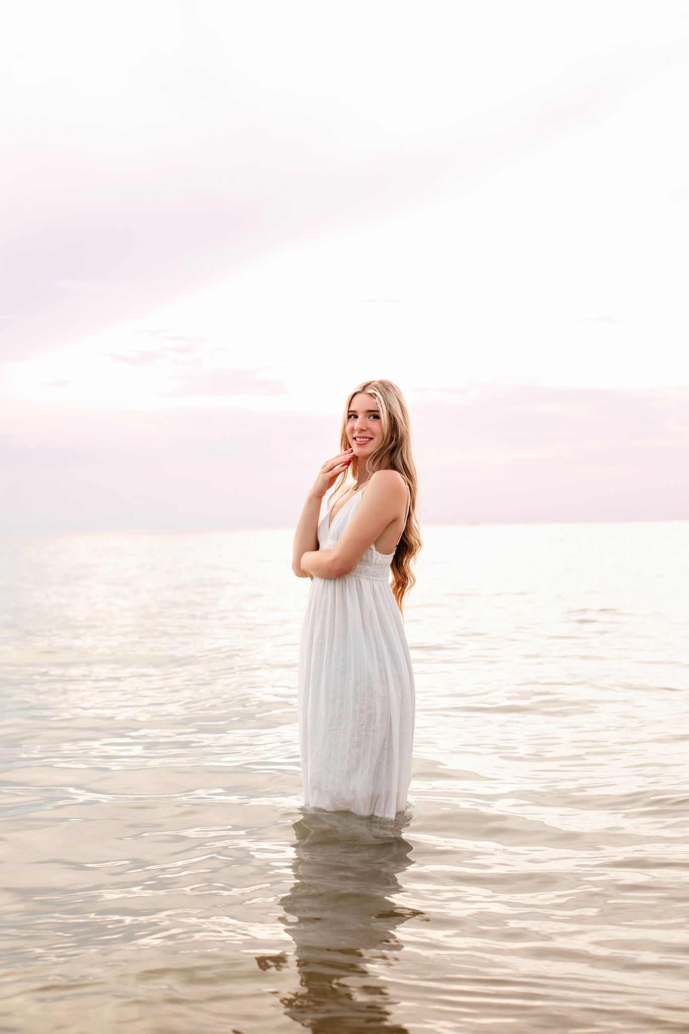 Young woman in white dress standing in calm ocean water at sunset, smiling over her shoulder.